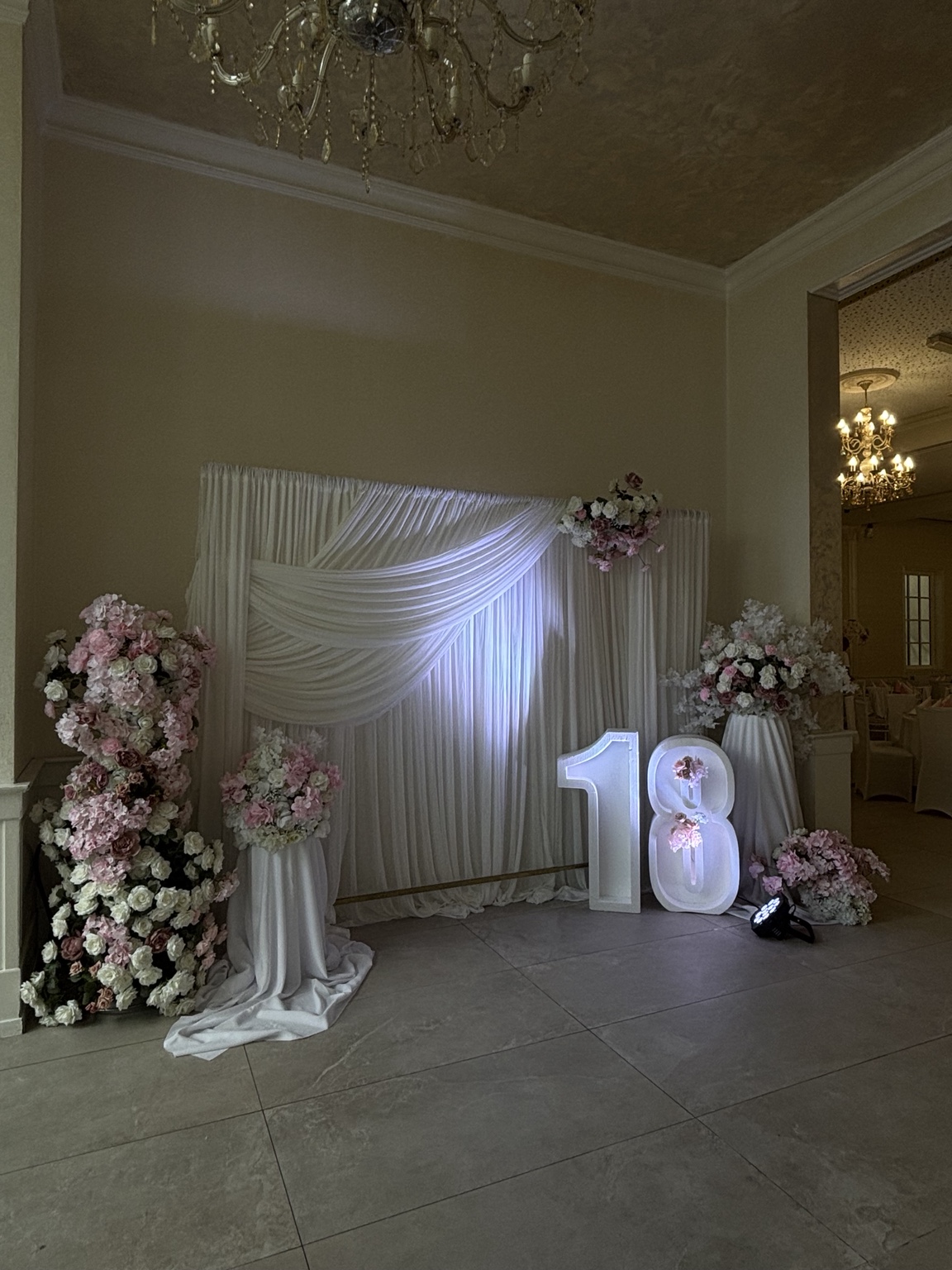 Weiße Stoffkulisse mit rosa Blumenarrangements, beleuchteter Zahl 18 und festlicher Lichteratmosphäre