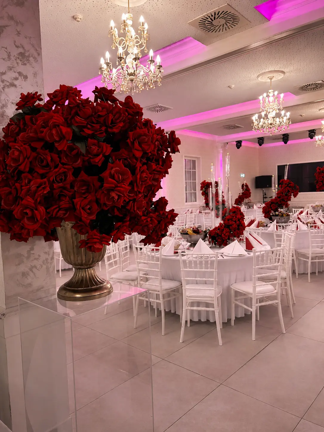 Festsaal mit üppigen roten Rosenarrangements, weißen Chiavari-Stühlen und Kristalllüstern in pinkem Licht