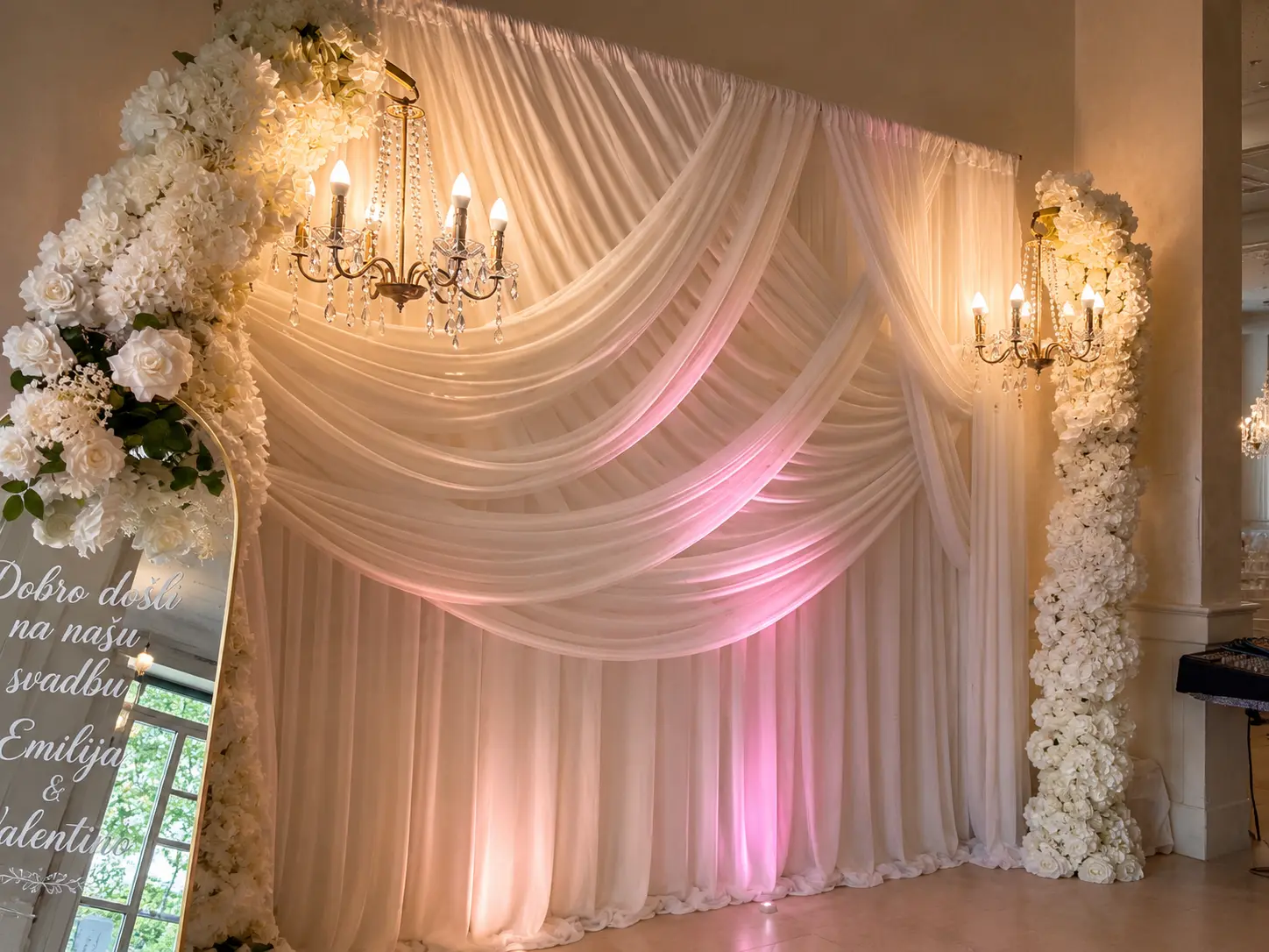Drapierte Hochzeitswand in Creme und Rosa mit Kristalllüstern, weißen Rosensäulen und Willkommensspiegel
