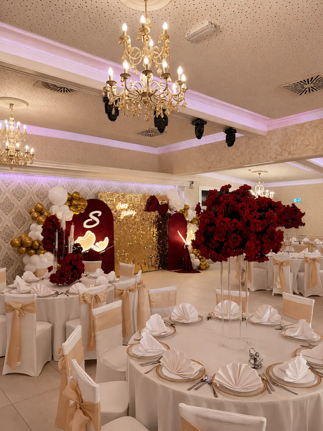 Eleganter Festsaal mit goldener Pailletten-Wand, roten Rosen, Gold-Ballongirlande und creme gedeckten Tischen