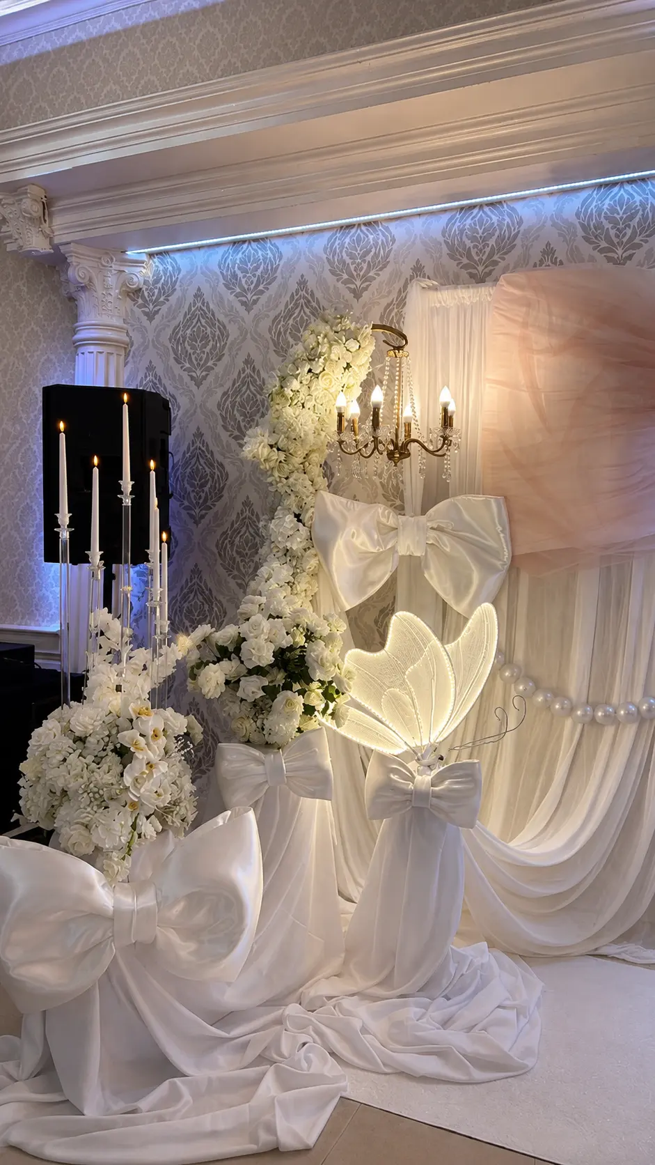 Elegante Fotowand in Weiß mit Satinschleifen, weißen Blumen, Kerzenleuchtern und beleuchtetem Schmetterling