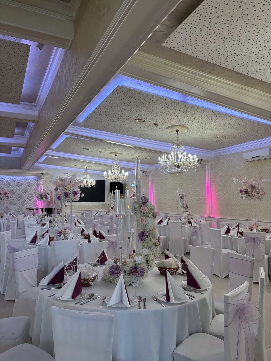 Eleganter Festsaal in Rosa und Lila mit hohen Blumenarrangements, Kronleuchtern und gedeckten Tischen