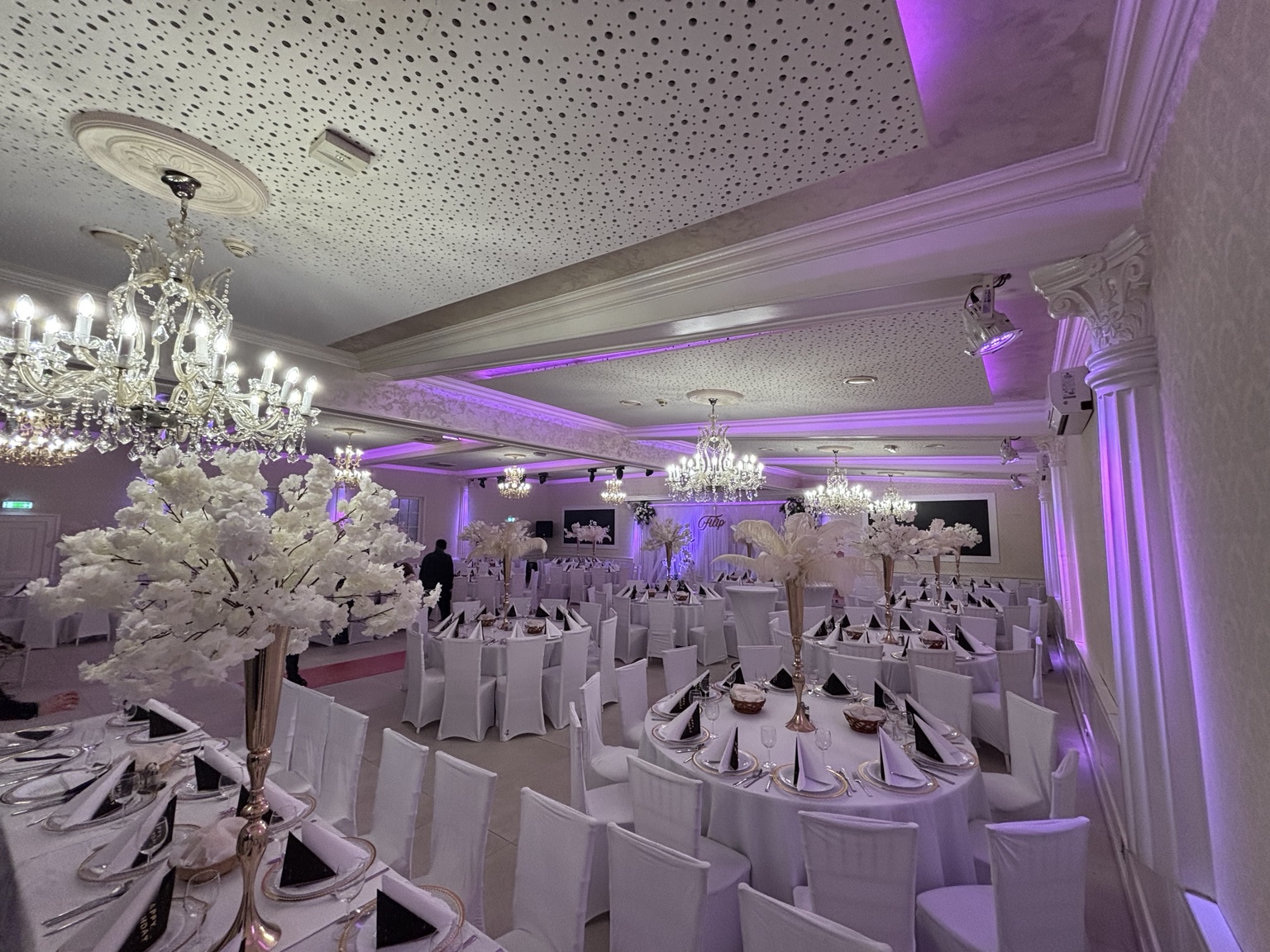 Festsaal in lila Licht mit weißen Blumenbäumen, gedeckten Tischen und prächtigen Kristalllüstern